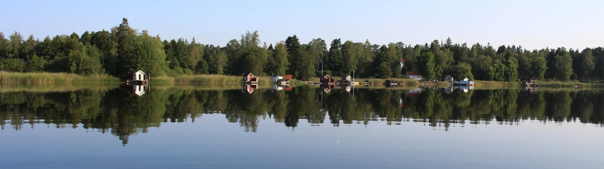 Sjön ligger spegelblank med skogsklädd strand och små byggnader längs vattenlinjen.